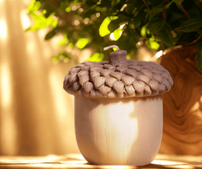 Realistic acorn-shaped candle with textured brown cap and smooth beige base, lit by warm sunlight with green foliage in the background.