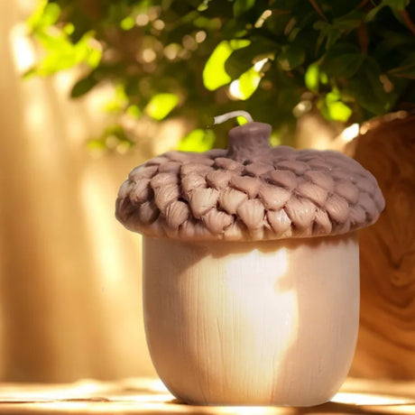 Realistic acorn-shaped candle with textured brown cap and smooth beige base, lit by warm sunlight with green foliage in the background.