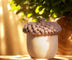 Realistic acorn-shaped candle with textured brown cap and smooth beige base, lit by warm sunlight with green foliage in the background.