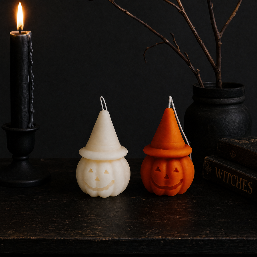 Two Halloween-themed wax candles on a dark wooden surface, one shaped like a cream-colored witch hat pumpkin and the other like an orange jack-o'-lantern with a pointed hat, styled with a lit black candle, a black cauldron, and books in the background. Pumpkin Hat Soy Wax Candles