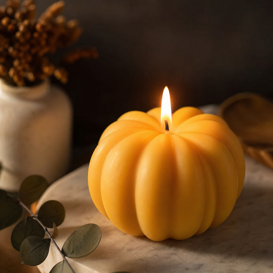 A yellow-orange pumpkin-shaped candle with a lit flame, styled on a marble surface with eucalyptus leaves and a ceramic vase in the background. Handmade Pumpkin Candle by Votive Candle Co