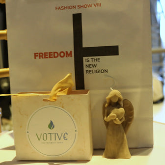 Votive candle with angel figurine in front of a 'Fashion Show VIII' sign.