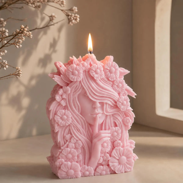 A pink sculpted flower goddess candle with a face and floral details, lit with a visible flame, set on a tabletop against a softly lit neutral background with dried flowers in the corner.