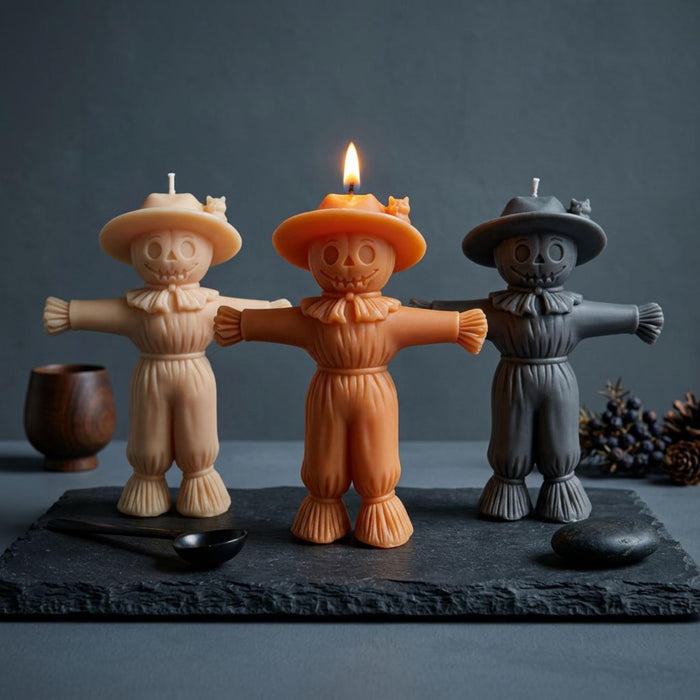 Three Halloween-themed scarecrow candles displayed on a dark slate board. The left candle is a light beige/cream color, the center candle is orange and is lit with a visible flame, and the right candle is dark gray. Scarecrow Soy Wax Candle – Halloween Fall Decor