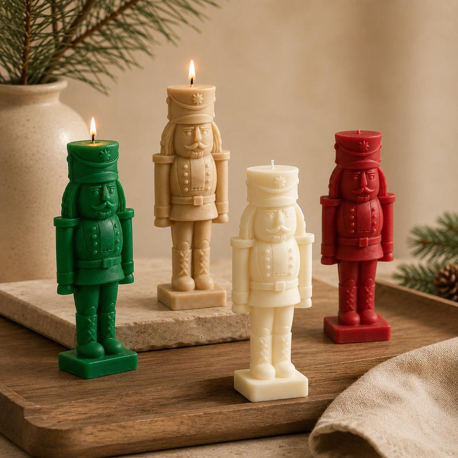 Four nutcracker-shaped candles in green, beige, cream, and red, arranged on a wooden tray with a warm neutral background and holiday greenery.Hand-Poured Nutcracker Candle: Sculptural Soy Wax Christmas Decor