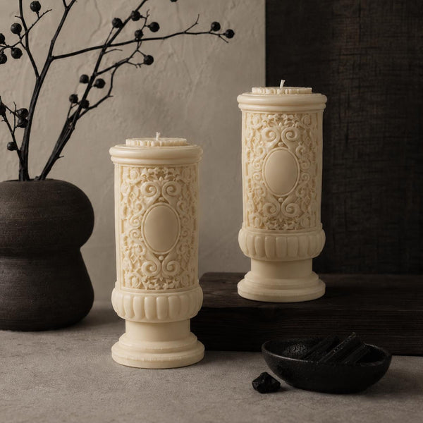 Gothic Pillar Candle, Dark Academia Decor, Victorian Pillar Candle. Two cream-colored pillar candles with ornate embossed scrollwork and oval medallions, styled on a dark wooden surface next to a dark ceramic vase with dried branches.