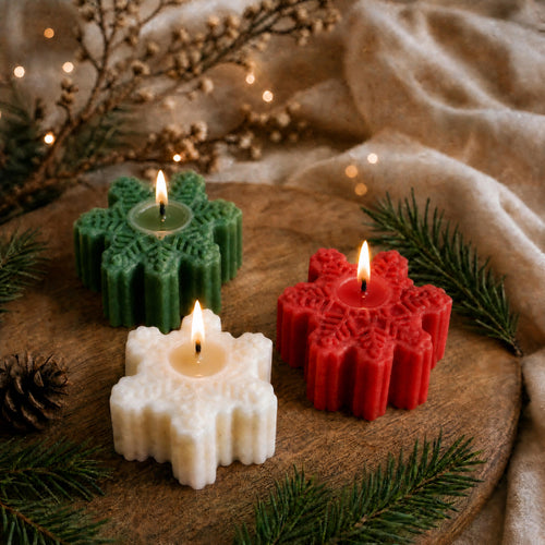 Three snowflake-shaped candle holders in green, red, and white, each holding a lit tealight candle, arranged on a rustic wooden board with evergreen branches, a pinecone, and soft beige fabric in the background.