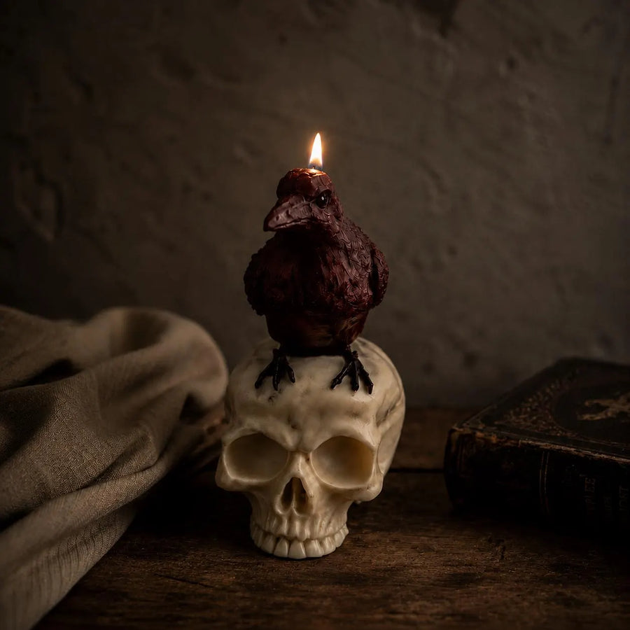 A dark brown crow-shaped candle with a lit flame sits on top of an off-white skull, placed on a rustic wooden surface with a draped cloth and an old book in a dim, moody setting. Raven Skull Candle – Large Handmade Gothic Sculptural Candle, Dark Academia Halloween Decor