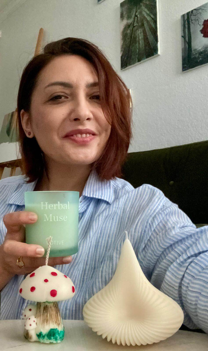 Woman holding a green candle labeled 'Herbal Musk' with decorative items on a table.