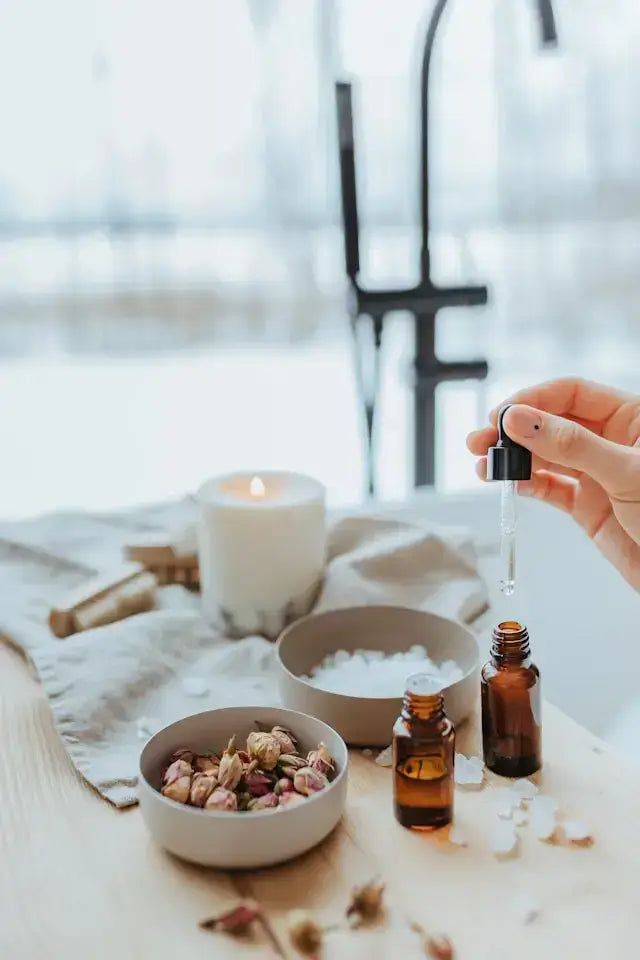 Hand adding drops from an essential oil bottle beside a candle and bath salts