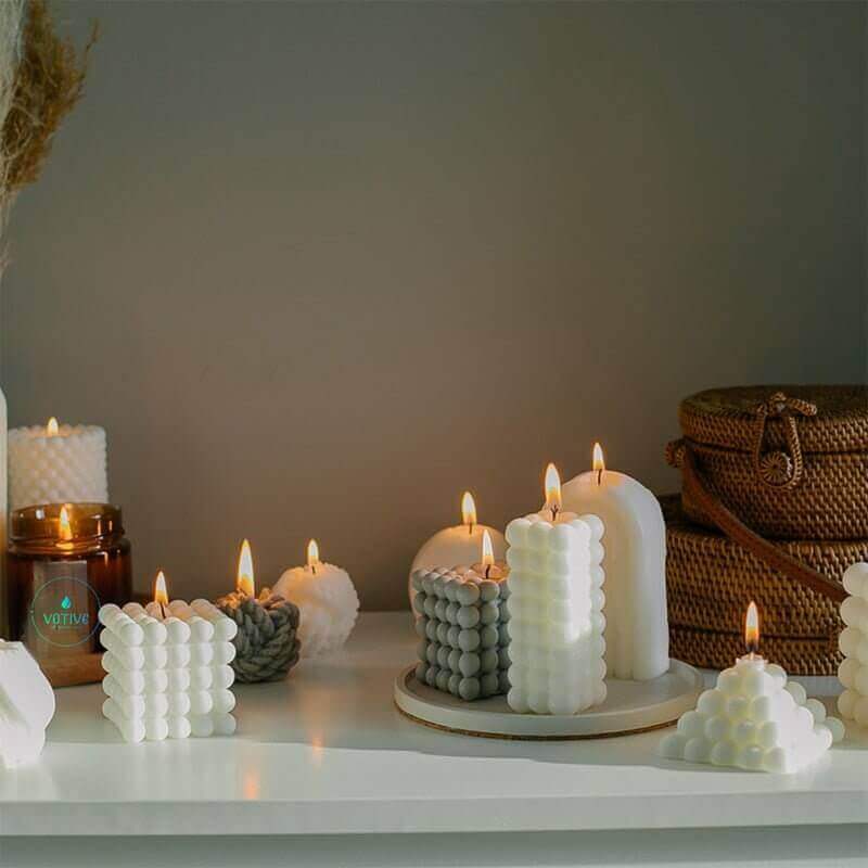 Lit bubble cube, pillar and pyramid candles on a white shelf with rattan baskets