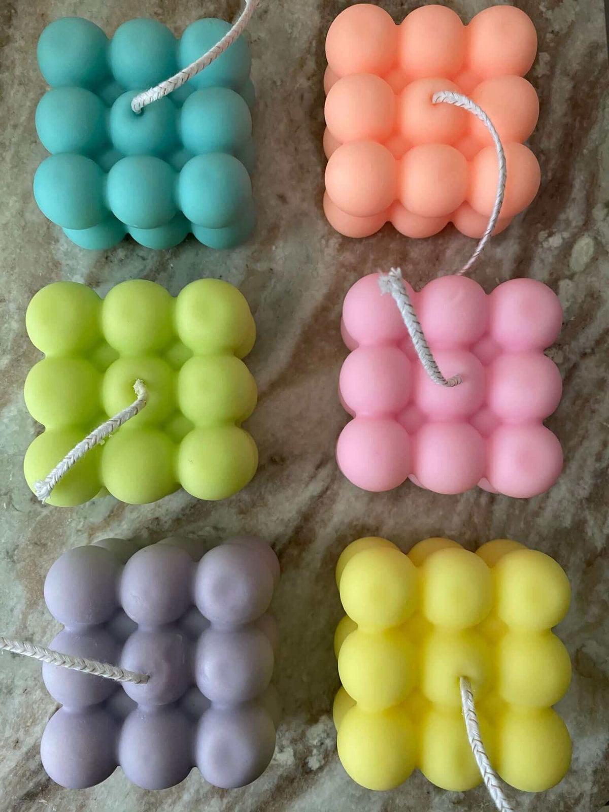 Six bubble cube candles in teal, peach, lime, pink, lavender and yellow on a sandy surface