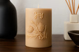 Beige candle with 'Joy' and leaf design on a wooden surface