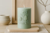Green candle with 'joy' and pine cone design on a wooden board with a neutral background