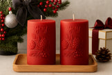 Two red candles with 'joy' and berry designs on a wooden tray, surrounded by Christmas decorations.