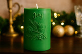 Green candle with 'Joy' and leaf design on a wooden surface with festive decorations.