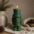 Nutcracker Christmas Candle — Large Sculptural Soy Wax Holiday Decor. 
A green nutcracker-shaped candle with a lit flame, styled on a beige cloth with pinecones and a ceramic vase in the background.