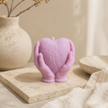 A lavender-pink heart-shaped candle held in two sculpted hands, decorated with embossed swirl patterns. The candle is styled on a textured stone slab next to a beige ceramic vase with dried branches and a neutral fabric background.