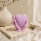 A lavender-pink heart-shaped candle held in two sculpted hands, decorated with embossed swirl patterns. The candle is styled on a textured stone slab next to a beige ceramic vase with dried branches and a neutral fabric background.