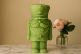 Green nutcracker candle on a marble surface with a vase of flowers in the background.
