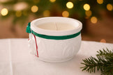 White ceramic mug with a candle inside, tied with a green ribbon, on a white surface with a blurred festive background.