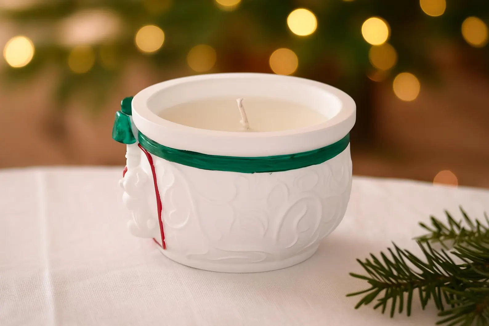 White ceramic mug with a candle inside, tied with a green ribbon, on a white surface with a blurred festive background.