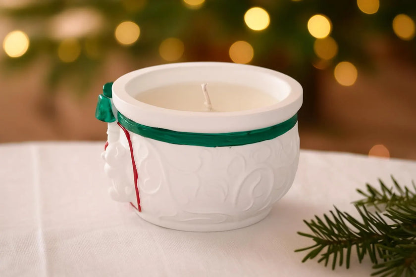 White ceramic mug with a candle inside, tied with a green ribbon, on a white surface with a blurred festive background.