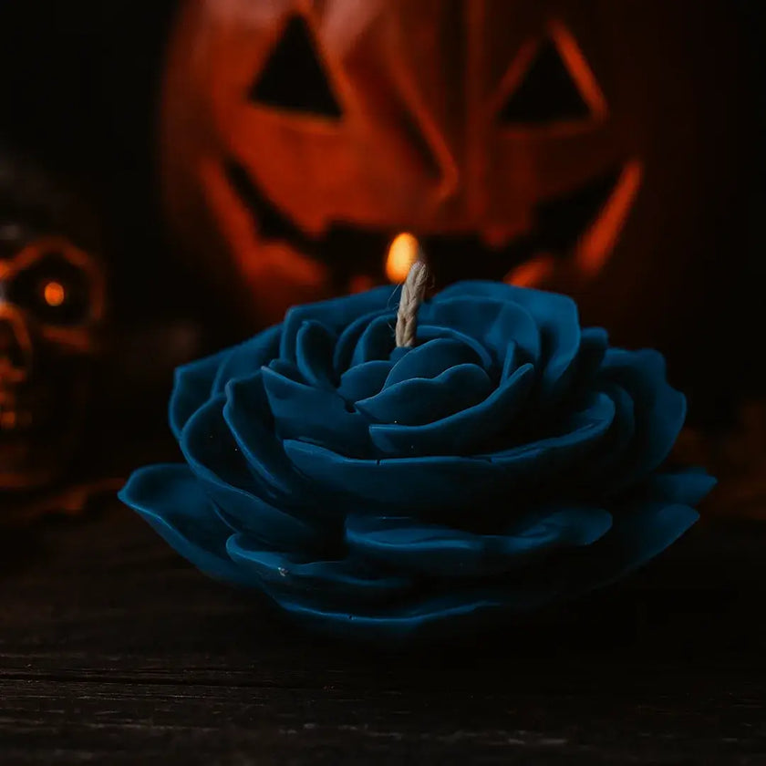 Black rose-shaped candle with a lit wick in front of a blurred jack-o'-lantern.