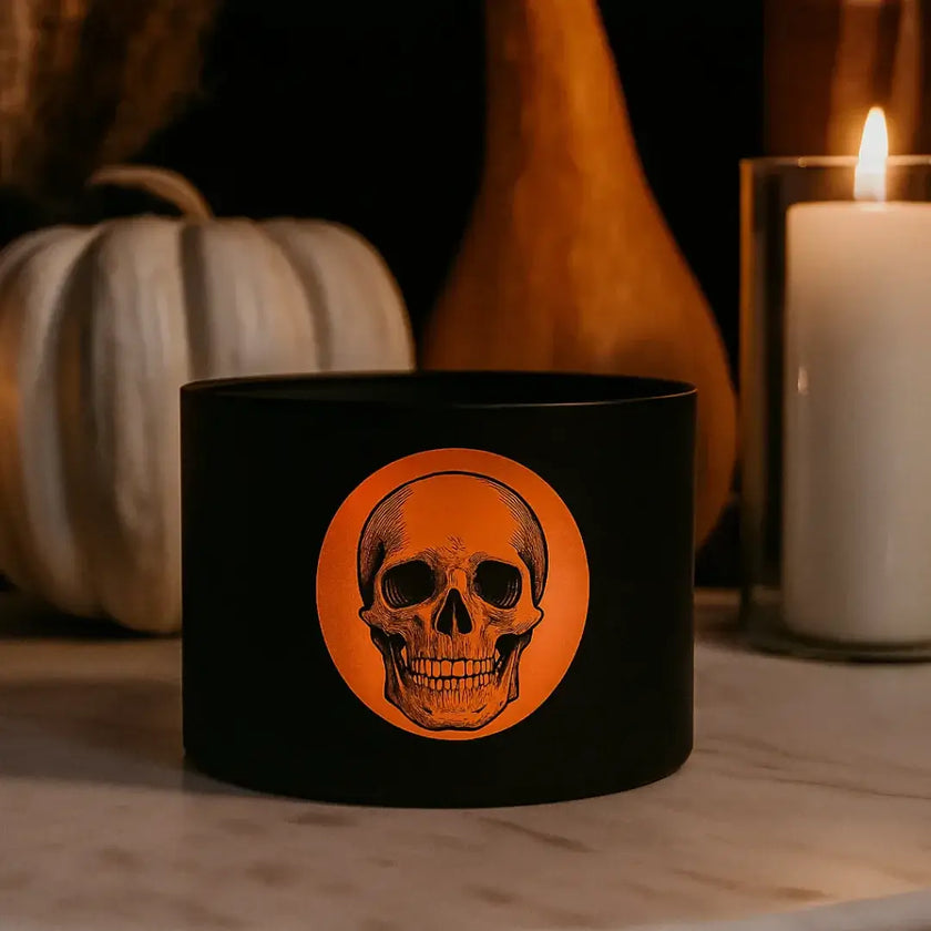 Black tin candle with a skull design on a wooden surface with pumpkins and a candle in the background
