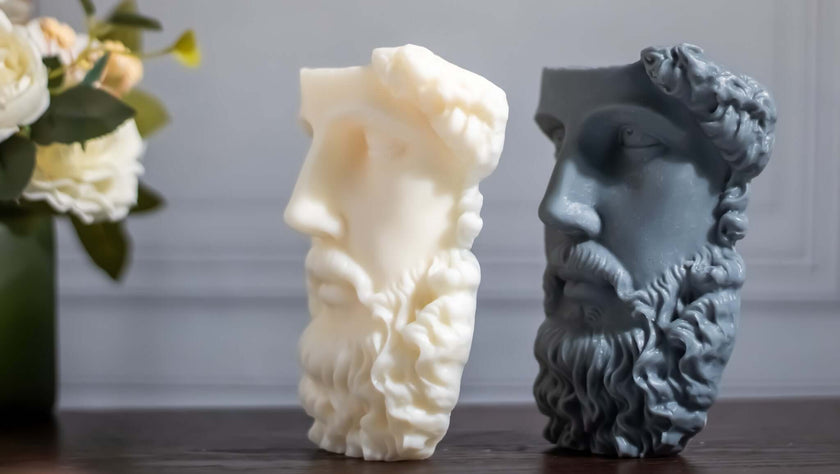 Sculptural soy wax candles in Greek face designs, one white and one dark gray, displayed with flowers.