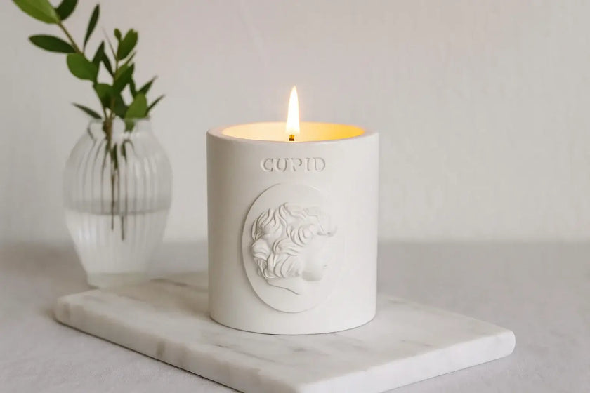 White candle labeled 'Cupid' on a marble coaster with a vase of greenery in the background