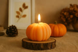 Pumpkin-shaped candle with a lit wick on a wooden stand, surrounded by pumpkins and pinecones. Mini pumpkin candle crafted from eco soy wax for Thanksgiving table centerpiece