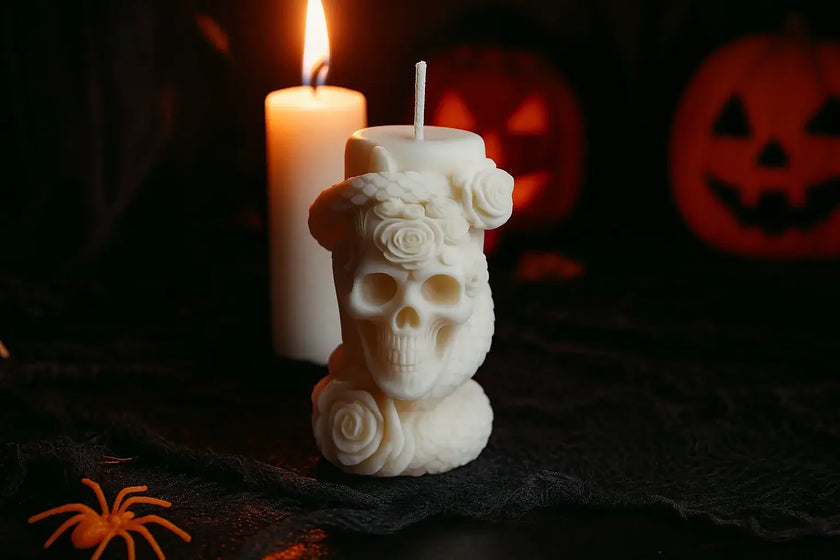 Candle shaped like a skull with roses, lit candle, and pumpkins in the background on a dark surface.