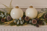 Decorative Christmas mantel with white ornaments, greenery, and pinecones.
