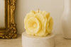 Decorative candle shaped like a flower on a marble base with a gold frame and white vase in the background.