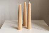 Three tall, thin, beige candles on a white surface with a neutral background