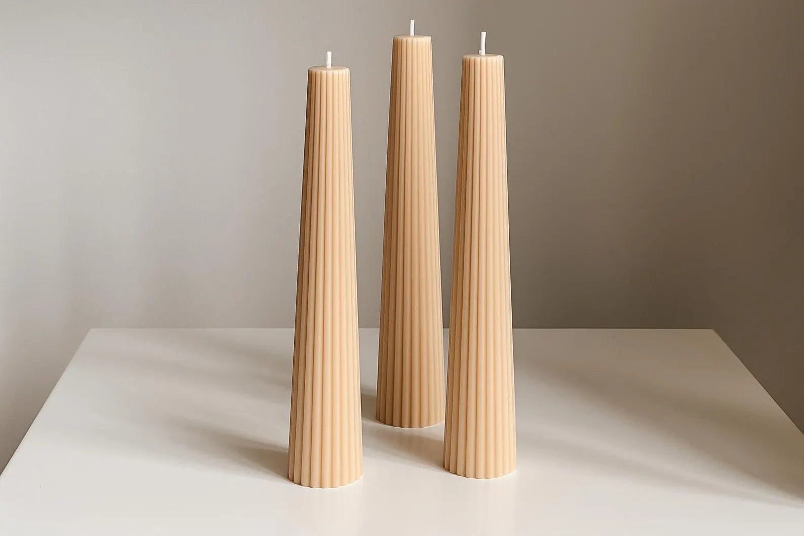 Three tall, thin, beige candles on a white surface with a neutral background