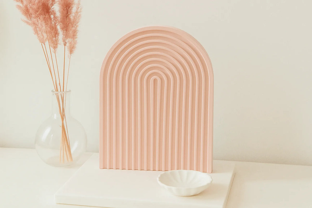 Decorative pink arch on a white surface with a vase of dried plants and a white bowl. - lit candle