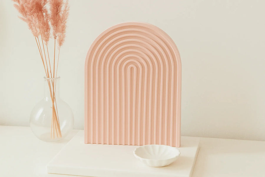 Decorative pink arch on a white surface with a vase of dried plants and a white bowl. - lit candle