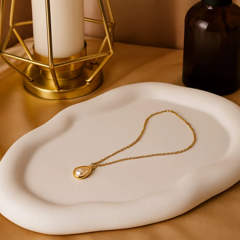 Gold necklace with a pendant on a white dish, with a candle and bottle in the background.