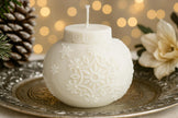 Decorative white spherical candle with snowflake patterns on a metallic plate with blurred lights and floral elements in the background.