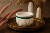 White candle in a decorative holder with green accent on a gold tray, surrounded by dried plants.