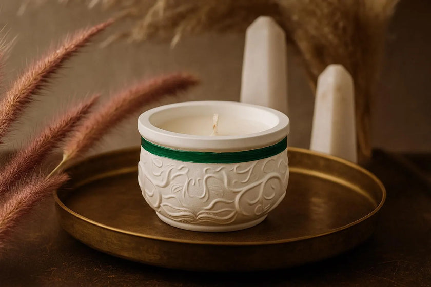 White candle in a decorative holder with green accent on a gold tray, surrounded by dried plants.