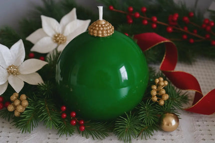 Green Christmas ornament with gold top surrounded by festive decorations including red berries, white flowers, and a red ribbon.