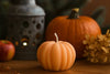 Small pumpkin-shaped candle with a larger pumpkin, apple, and lantern in the background on a wooden surface. Halloween pumpkin candle perfect for autumn decorations and seasonal gifts