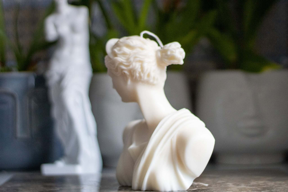 Hand-poured Greek-inspired Venus bust candle in white soy wax, with a classical statue softly blurred behind it