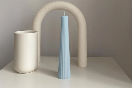 Light blue textured candle on a white surface with a beige arch in the background