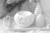 White ceramic candle holder with a lit candle on a snowy surface with decorative trees and pine cones.