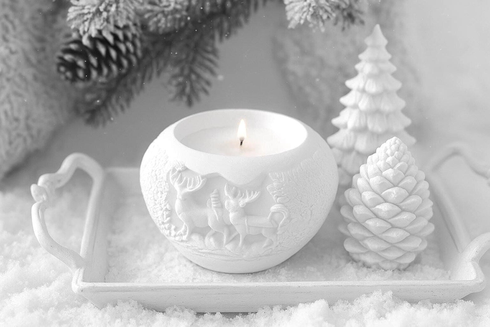 White ceramic candle holder with a lit candle on a snowy surface with decorative trees and pine cones.