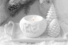 White ceramic candle holder with a lit candle on a snowy surface with decorative trees and pine cones.